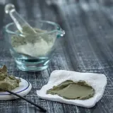 Pâte d'argile verte prête à l'emploi, avec une cuillère sur une petite assiette et une autre portion sur un tissu blanc. Un mortier en verre rempli de poudre d'argile est visible en arrière-plan.