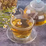 Une tasse de thé à la camomille matricaire, une théière en verre et des fleurs fraîches sur fond texturé.