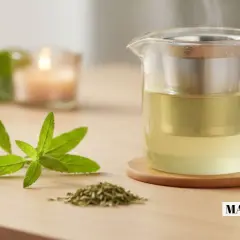 Une théière en verre fumante contient une tisane verveine dorée. Des feuilles fraîches et séchées de verveine, ainsi qu'une tranche de citron, complètent la scène.