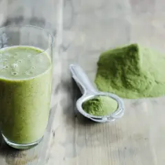 Un verre de smoothie vert et une cuillère remplie de poudre d'argile verte à boire sur une table en bois.