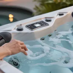 Main d'une personne effleurant l'eau bouillonnante d'un **dessous spa** moderne, avec des jets visibles et un panneau de contrôle.