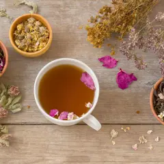 Tasses de thé aux herbes et pétales de rose, idéales pour apaiser la nausée. Des herbes séchées et des fleurs décorent la table en bois.