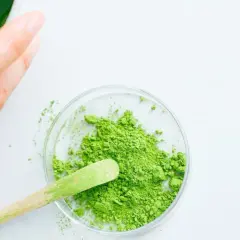 Une main prépare du matcha. Une cuillère en bois prélève la poudre verte d'une coupelle, suggérant la quantité de matcha par tasse.