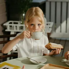 Une petite fille blonde boit du thé, se demandant à quel âge on peut boire du thé. Elle tient un éclair à la main.