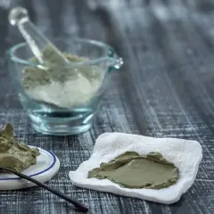 Pâte d'argile verte prête à l'emploi, avec une cuillère sur une petite assiette et une autre portion sur un tissu blanc. Un mortier en verre rempli de poudre d'argile est visible en arrière-plan.