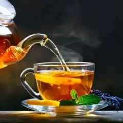 Un thé ambré, aux reflets de la noire, est versé d'une théière en verre dans une tasse ornée de citron et de menthe.