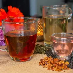 Tasses de thé fumant, un pichet de thé clair et une petite tasse de thé rose. Des fleurs et des feuilles de thé séchées complètent cette scène de dégustation de noms de thé.