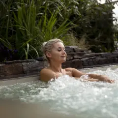 Femme détendue dans un jacuzzi, ignorant les dangers du spa. L'eau bouillonnante et la végétation luxuriante créent une scène paisible, mais attention aux infections.