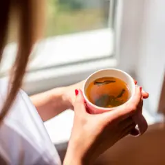 Une femme aux ongles rouges tient une tasse de thé fumant, un rituel quotidien pour se détendre près de la fenêtre.