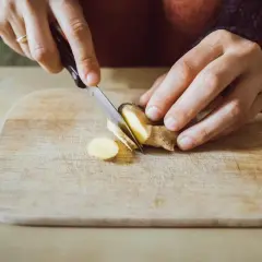Préparation d'un thé au gingembre le soir. Des mains coupent finement du gingembre sur une planche en bois.