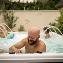 Un homme barbu rit en ajustant les commandes d'un spa jacuzzi. Sa famille profite des jets d'eau.