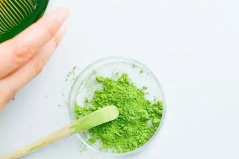 Une main prépare du matcha. Une cuillère en bois prélève la poudre verte d'une coupelle, suggérant la quantité de matcha par tasse.