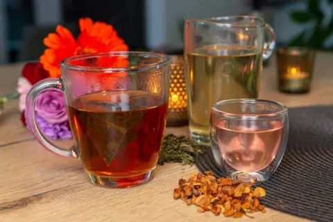 Tasses de thé fumant, un pichet de thé clair et une petite tasse de thé rose. Des fleurs et des feuilles de thé séchées complètent cette scène de dégustation de noms de thé.