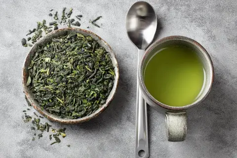 Thé vert en vrac dans un bol, une tasse de thé vert et une cuillère. Le thé vert est connu pour ses bienfaits, mais il peut aussi avoir des egcg effets secondaires.