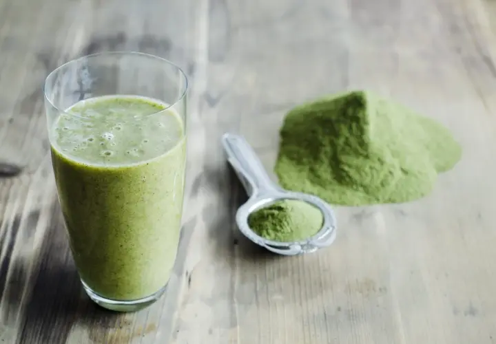 Un verre de smoothie vert et une cuillère remplie de poudre d'argile verte à boire sur une table en bois.