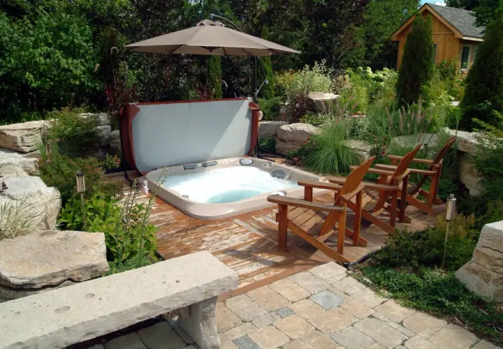Aménagement spa gonflable dans un jardin luxuriant avec des chaises en bois, un parasol et une petite cabane en bois.