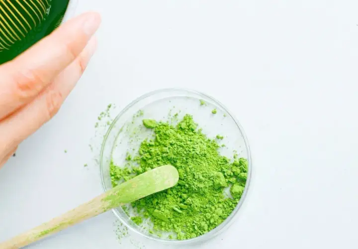 Une main prépare du matcha. Une cuillère en bois prélève la poudre verte d'une coupelle, suggérant la quantité de matcha par tasse.