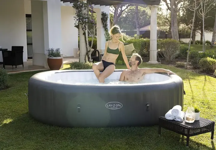 Couple profitant de la taille jacuzzi gonflable dans un jardin ensoleillé. Serviettes et boisson à proximité.