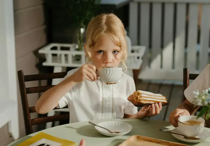 Une petite fille blonde boit du thé, se demandant à quel âge on peut boire du thé. Elle tient un éclair à la main.