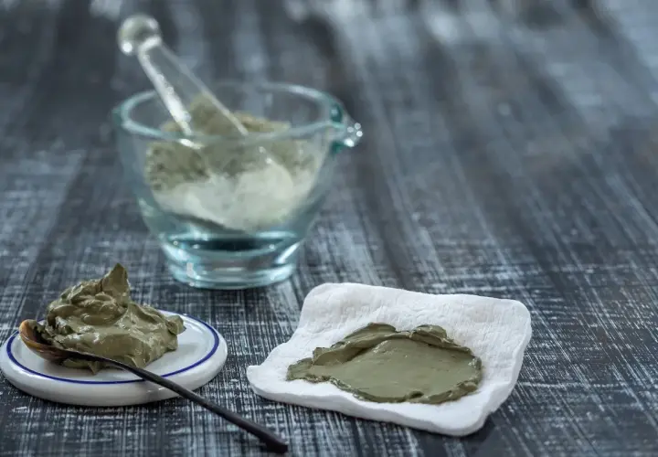 Pâte d'argile verte prête à l'emploi, avec une cuillère sur une petite assiette et une autre portion sur un tissu blanc. Un mortier en verre rempli de poudre d'argile est visible en arrière-plan.