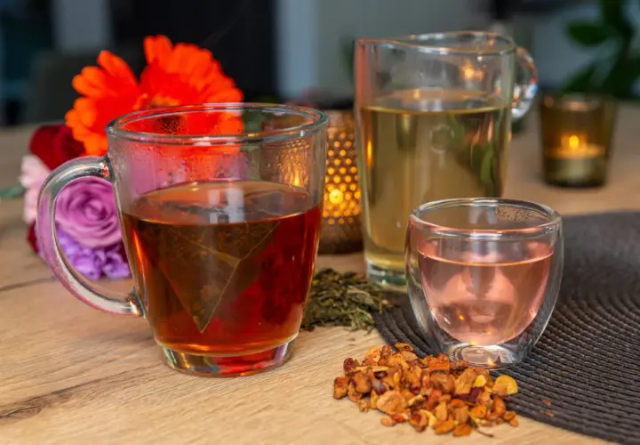 Tasses de thé fumant, un pichet de thé clair et une petite tasse de thé rose. Des fleurs et des feuilles de thé séchées complètent cette scène de dégustation de noms de thé.