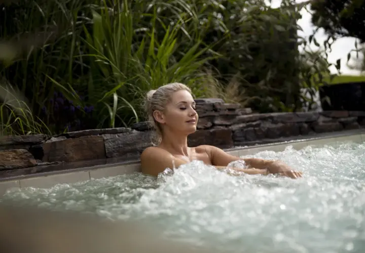 Femme détendue dans un jacuzzi, ignorant les dangers du spa. L'eau bouillonnante et la végétation luxuriante créent une scène paisible, mais attention aux infections.