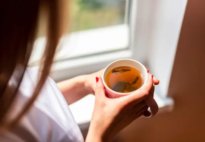 Une femme aux ongles rouges tient une tasse de thé fumant, un rituel quotidien pour se détendre près de la fenêtre.