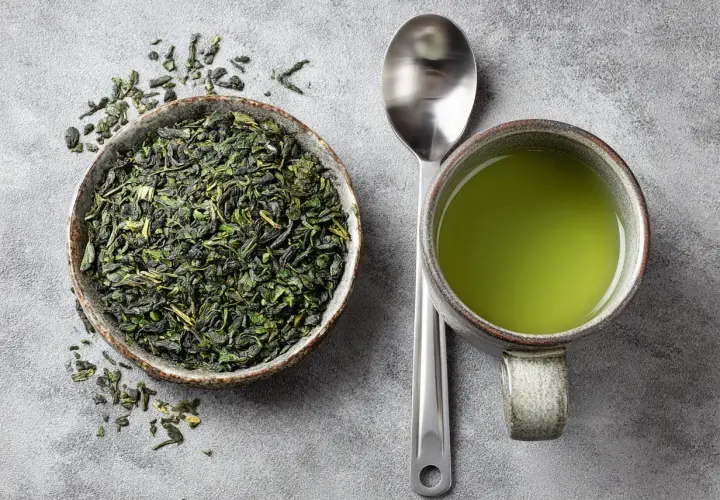 Thé vert en vrac dans un bol, une tasse de thé vert et une cuillère. Le thé vert est connu pour ses bienfaits, mais il peut aussi avoir des egcg effets secondaires.