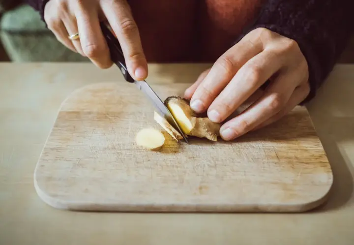 Préparation d'un thé au gingembre le soir. Des mains coupent finement du gingembre sur une planche en bois.