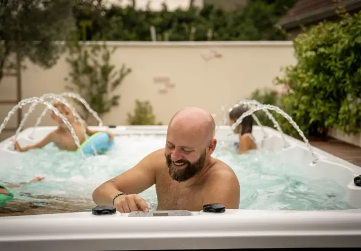 Un homme barbu rit en ajustant les commandes d'un spa jacuzzi. Sa famille profite des jets d'eau.