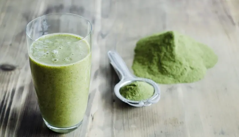 Un verre de smoothie vert et une cuillère remplie de poudre d'argile verte à boire sur une table en bois.