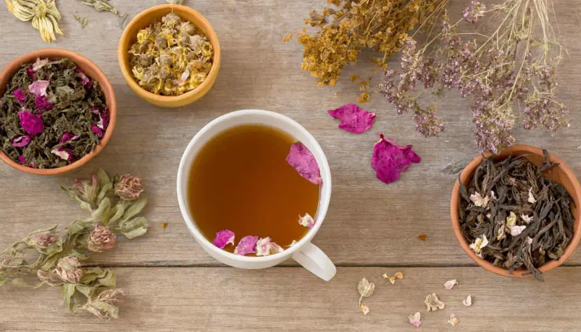 Tasses de thé aux herbes et pétales de rose, idéales pour apaiser la nausée. Des herbes séchées et des fleurs décorent la table en bois.