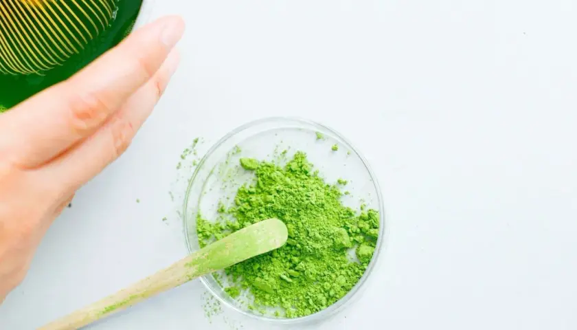 Une main prépare du matcha. Une cuillère en bois prélève la poudre verte d'une coupelle, suggérant la quantité de matcha par tasse.