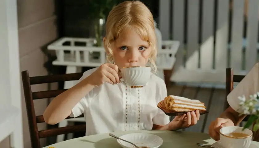 Une petite fille blonde boit du thé, se demandant à quel âge on peut boire du thé. Elle tient un éclair à la main.