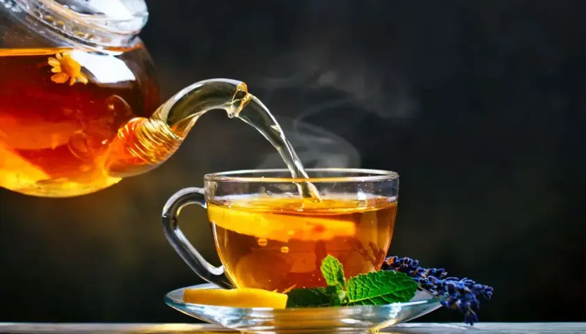 Un thé ambré, aux reflets de la noire, est versé d'une théière en verre dans une tasse ornée de citron et de menthe.