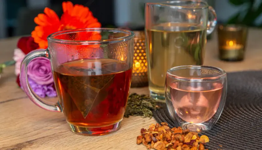 Tasses de thé fumant, un pichet de thé clair et une petite tasse de thé rose. Des fleurs et des feuilles de thé séchées complètent cette scène de dégustation de noms de thé.