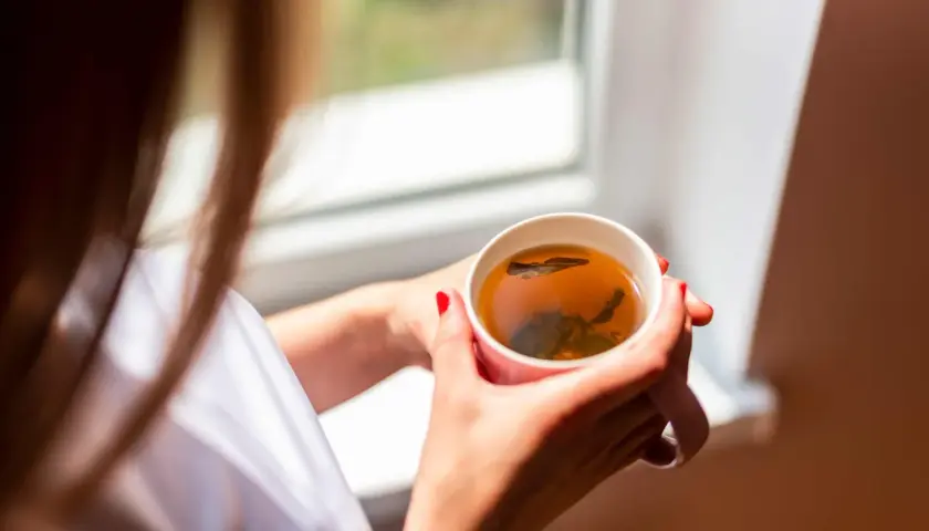 Une femme aux ongles rouges tient une tasse de thé fumant, un rituel quotidien pour se détendre près de la fenêtre.