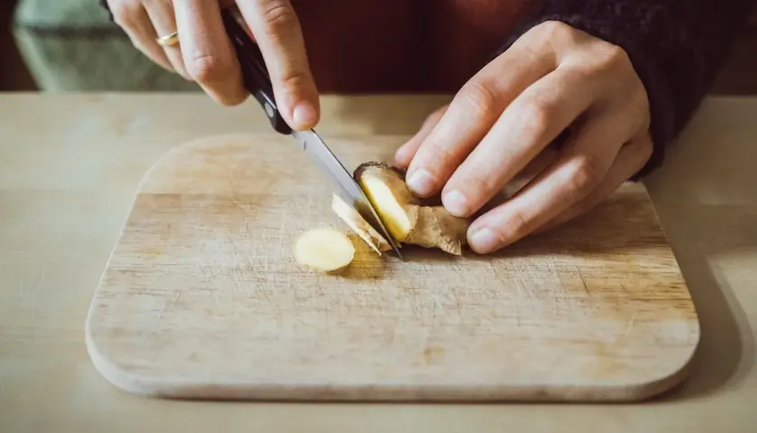Préparation d'un thé au gingembre le soir. Des mains coupent finement du gingembre sur une planche en bois.