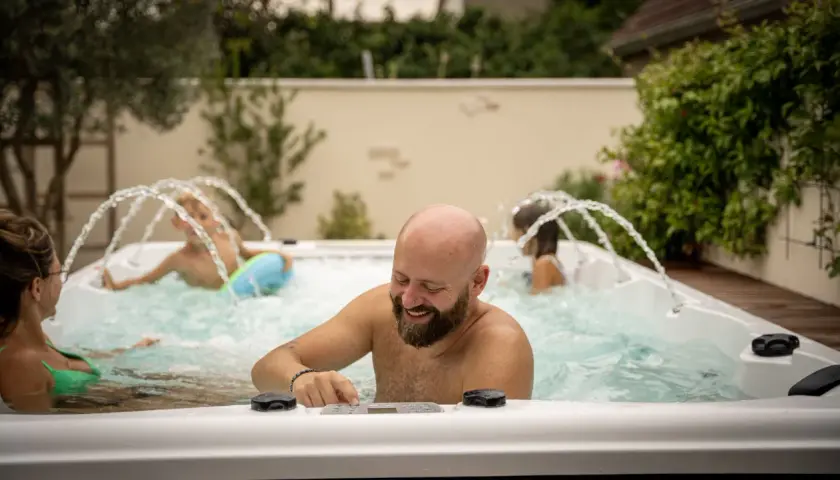 Un homme barbu rit en ajustant les commandes d'un spa jacuzzi. Sa famille profite des jets d'eau.