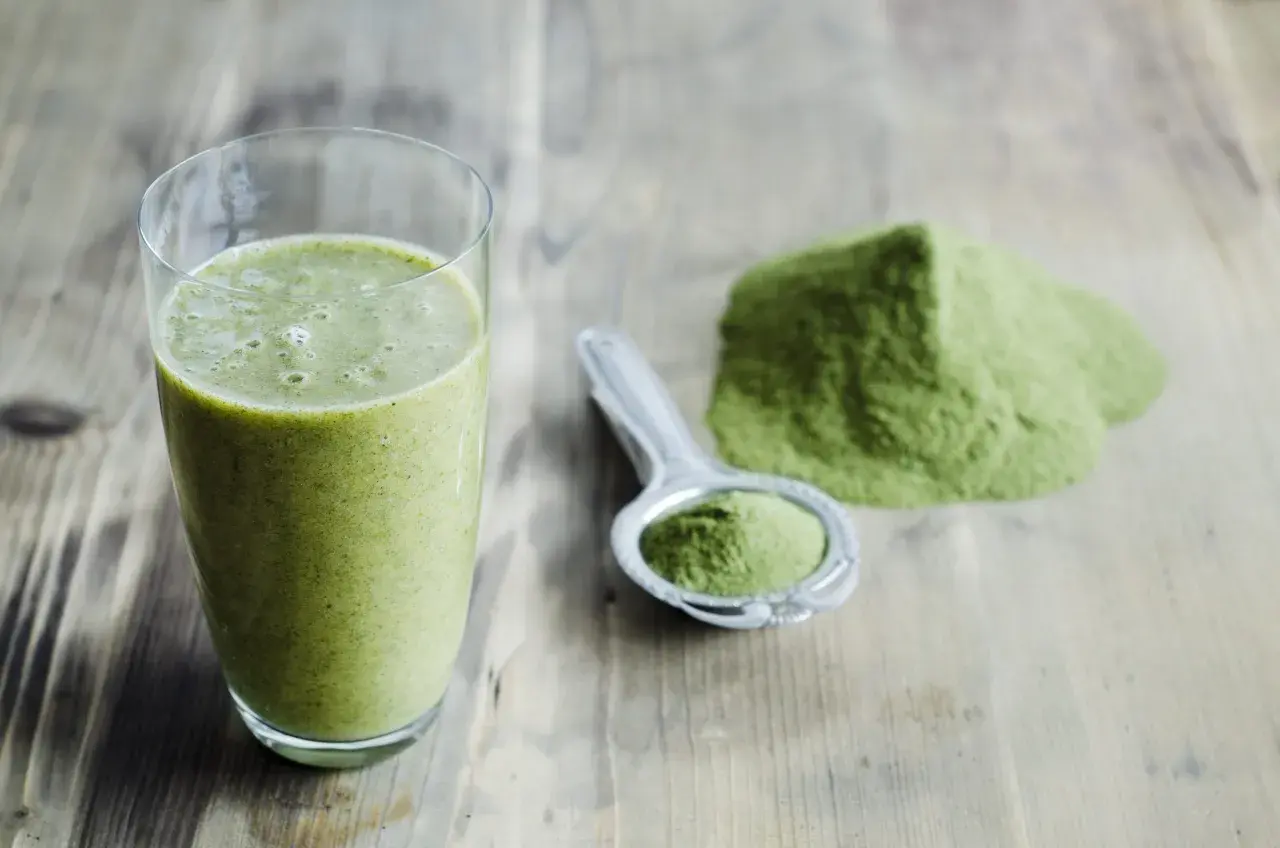 Un verre de smoothie vert et une cuillère remplie de poudre d'argile verte à boire sur une table en bois.