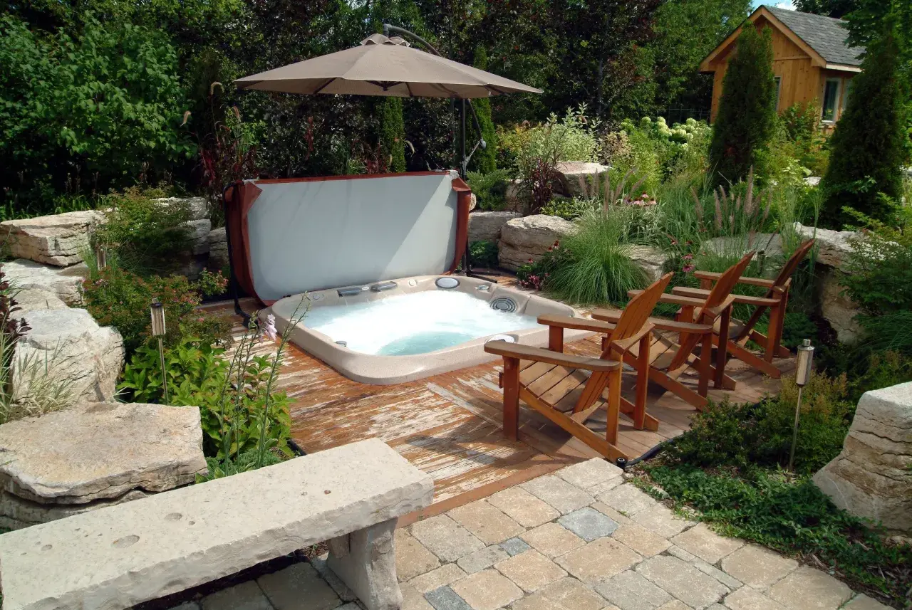 Aménagement spa gonflable dans un jardin luxuriant avec des chaises en bois, un parasol et une petite cabane en bois.