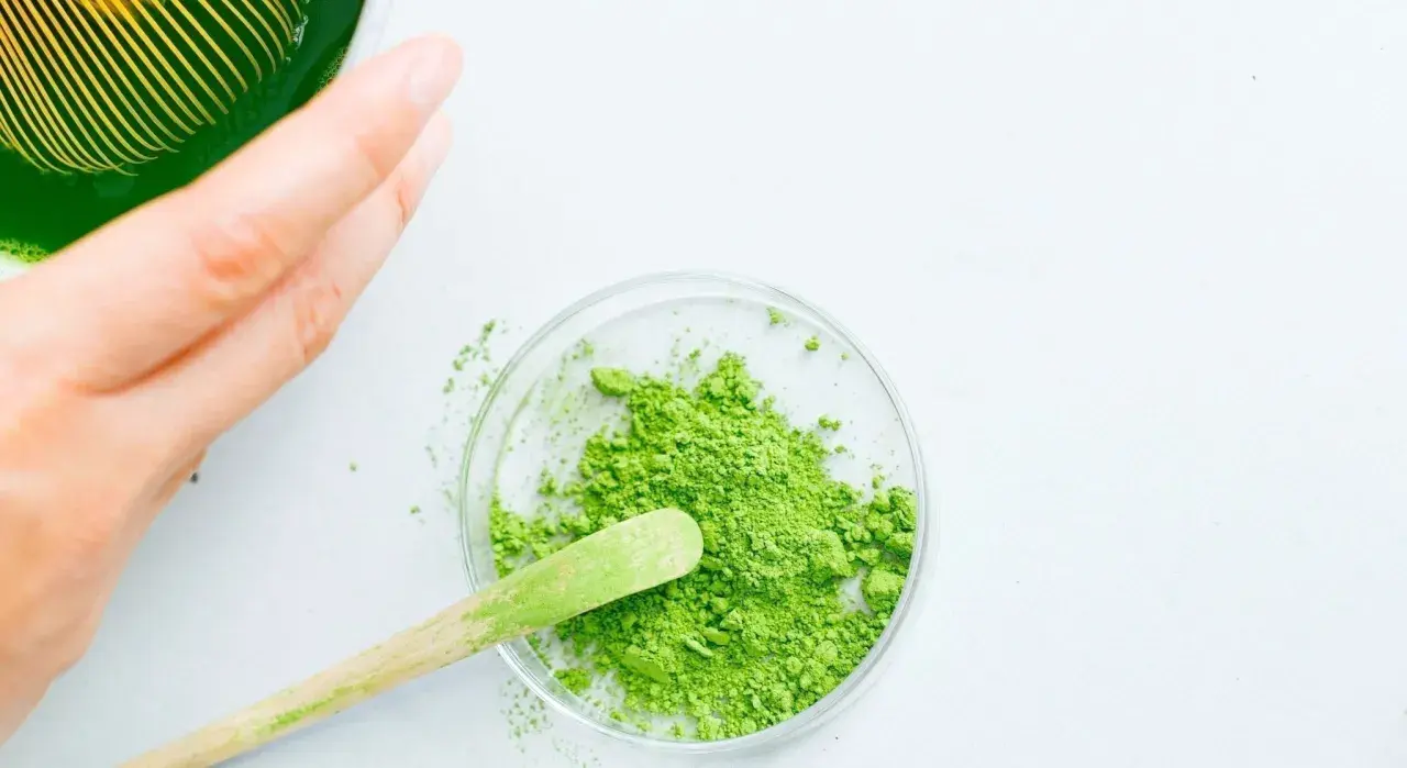 Une main prépare du matcha. Une cuillère en bois prélève la poudre verte d'une coupelle, suggérant la quantité de matcha par tasse.