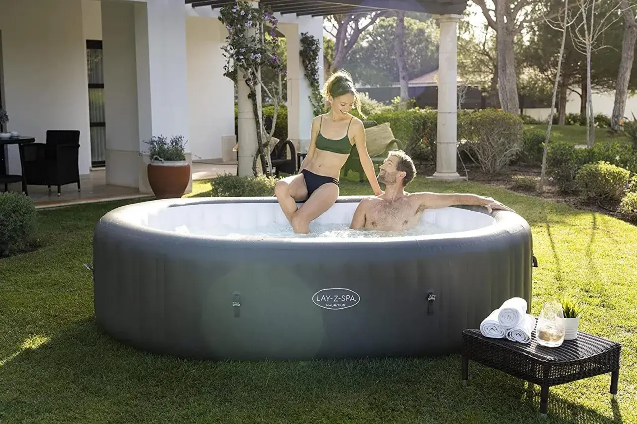 Couple profitant de la taille jacuzzi gonflable dans un jardin ensoleillé. Serviettes et boisson à proximité.