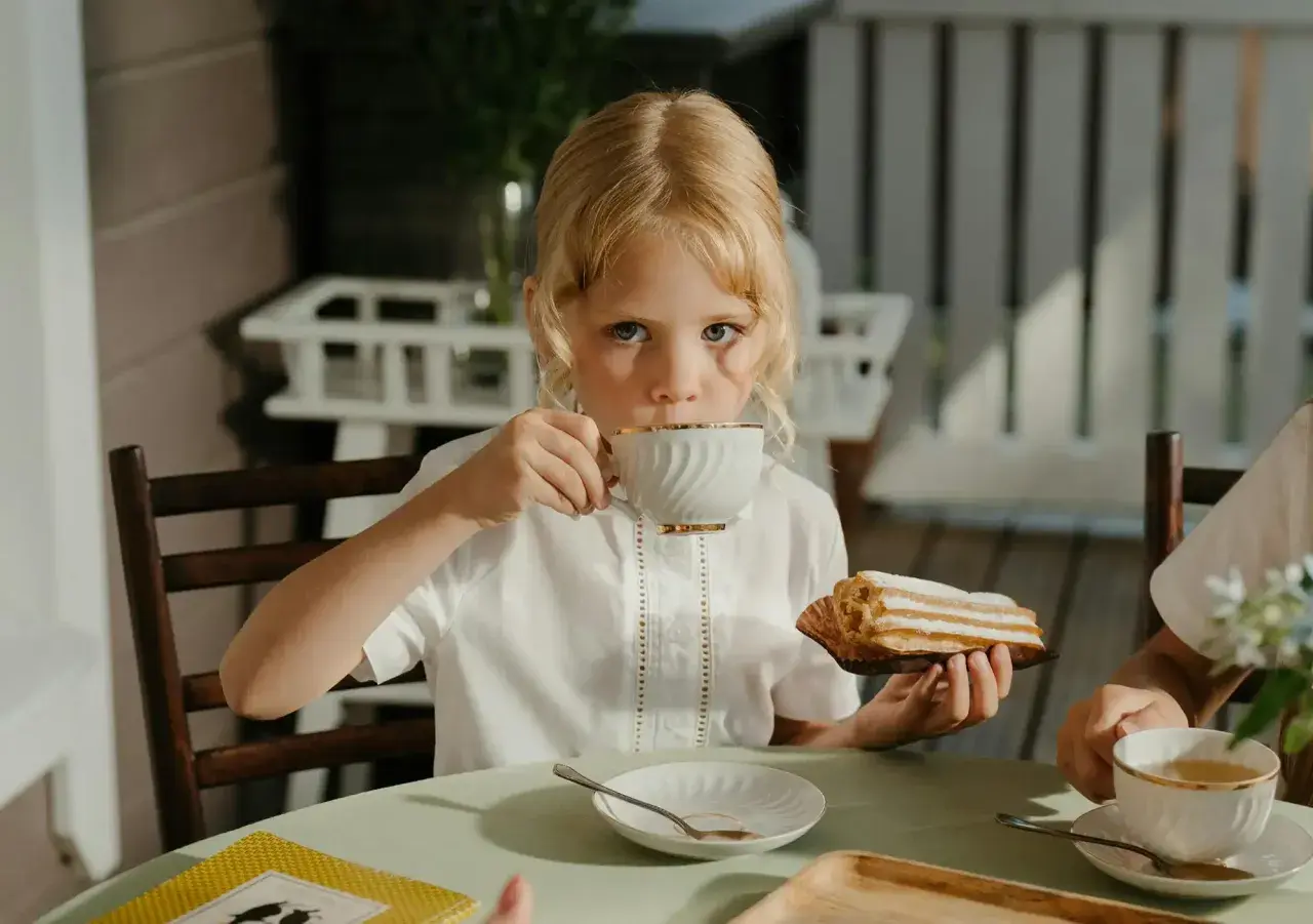 Une petite fille blonde boit du thé, se demandant à quel âge on peut boire du thé. Elle tient un éclair à la main.