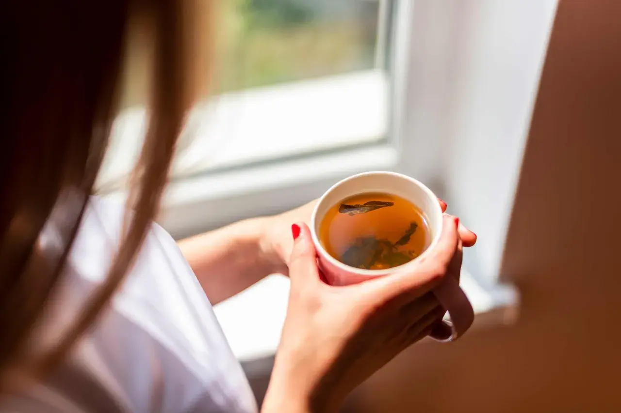 Une femme aux ongles rouges tient une tasse de thé fumant, un rituel quotidien pour se détendre près de la fenêtre.