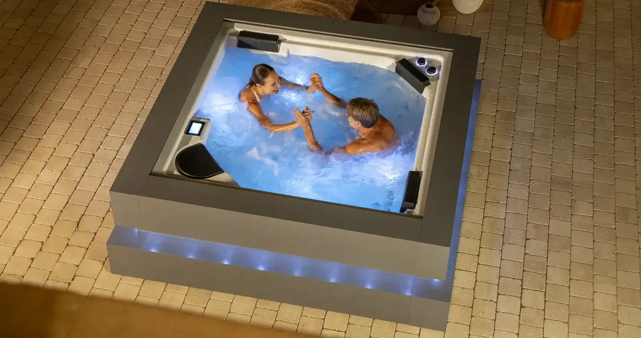 Un couple se tient la main dans un jacuzzi illuminé. Les jets massants promettent de soulager les courbatures après le jacuzzi.